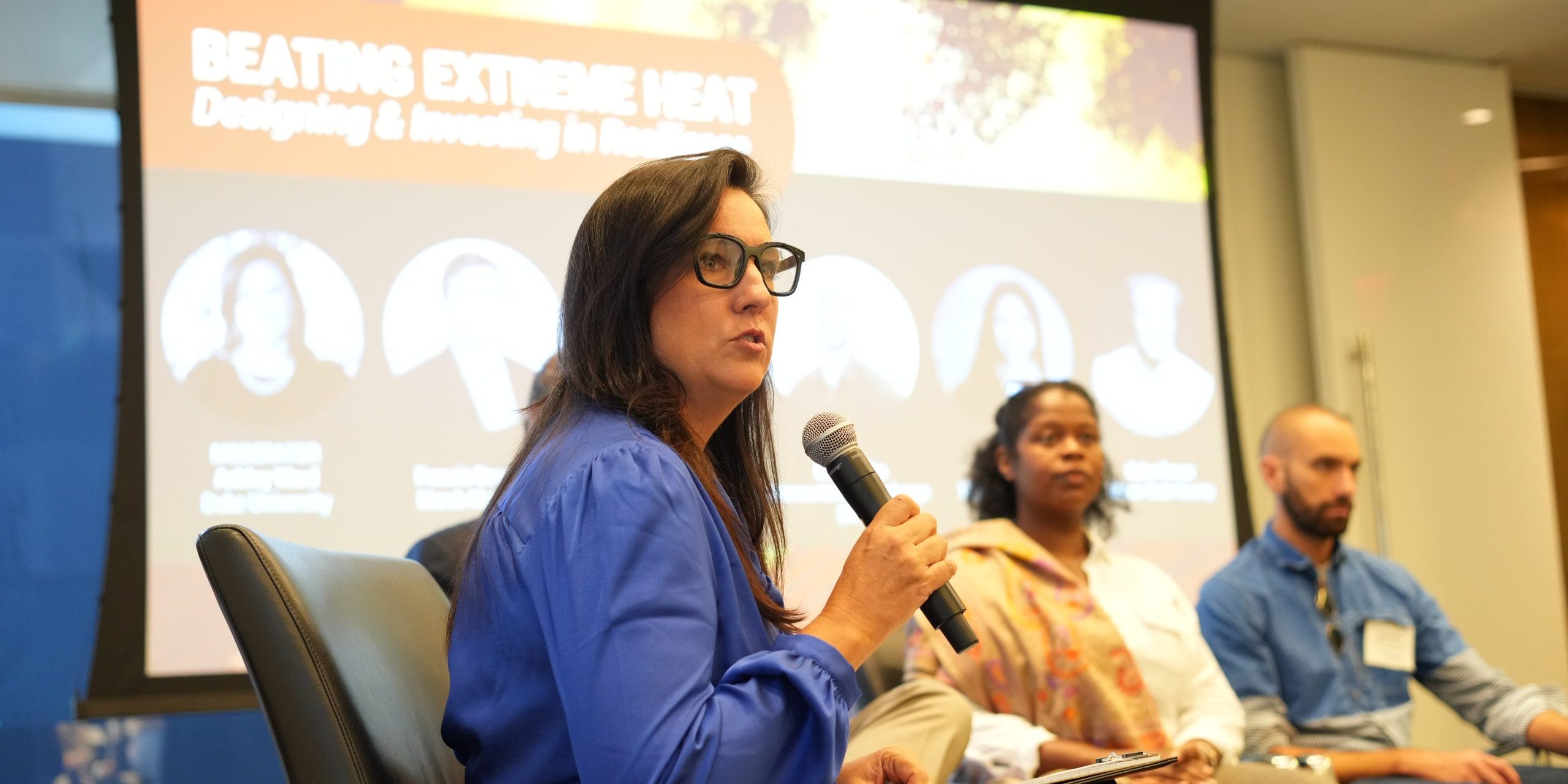 Panelist holds mic as two other panelists look on. In background is a slide that says Beating Extreme Heat.