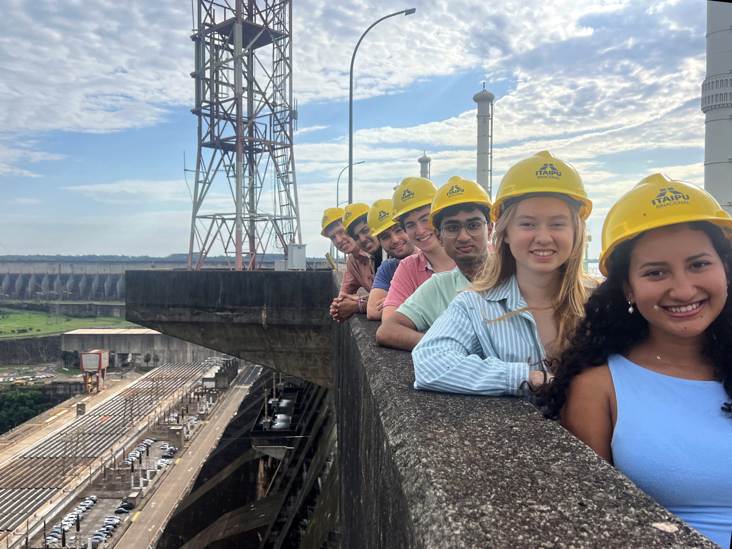 Students wearing hard hats as they stand against a concrete railing at Itaipu Dam, with dam infrastructure behind them.