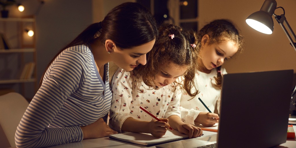 An adult and two children look at something on a table that is illuminated by a desk lamp